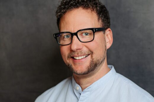 Smiling man with curly hair, black glasses, and a light blue shirt against a dark background.