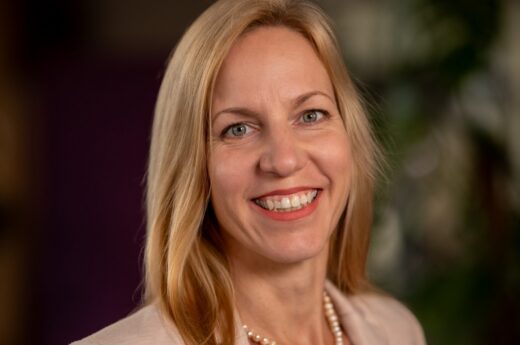 Smiling blonde woman wearing a beige blazer and pearl necklace, photographed in close-up.
