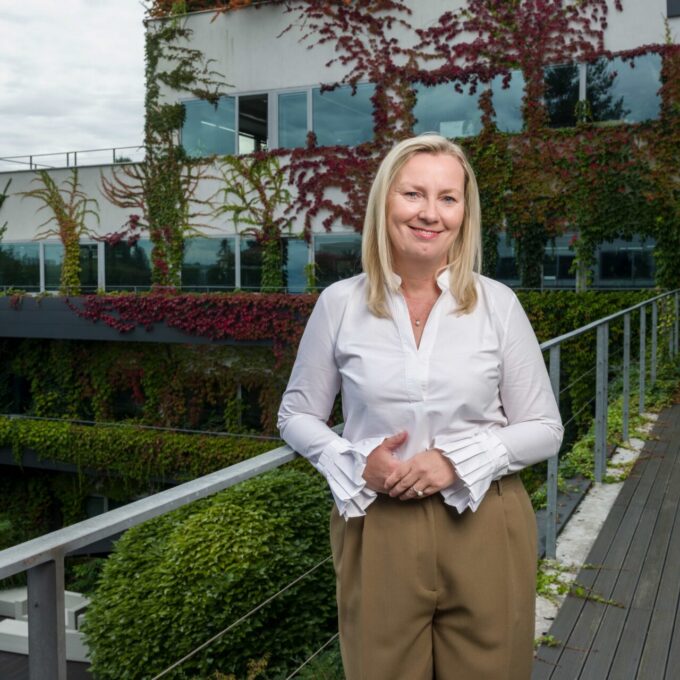 Blondýna v bílé halence stojí před moderní budovou porostlou zelení.